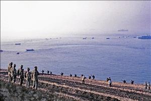 Men in uniform stand on a bluff facing towards the sea. At the water's edge, men run with guns. Warships of various sizes are off shore. 